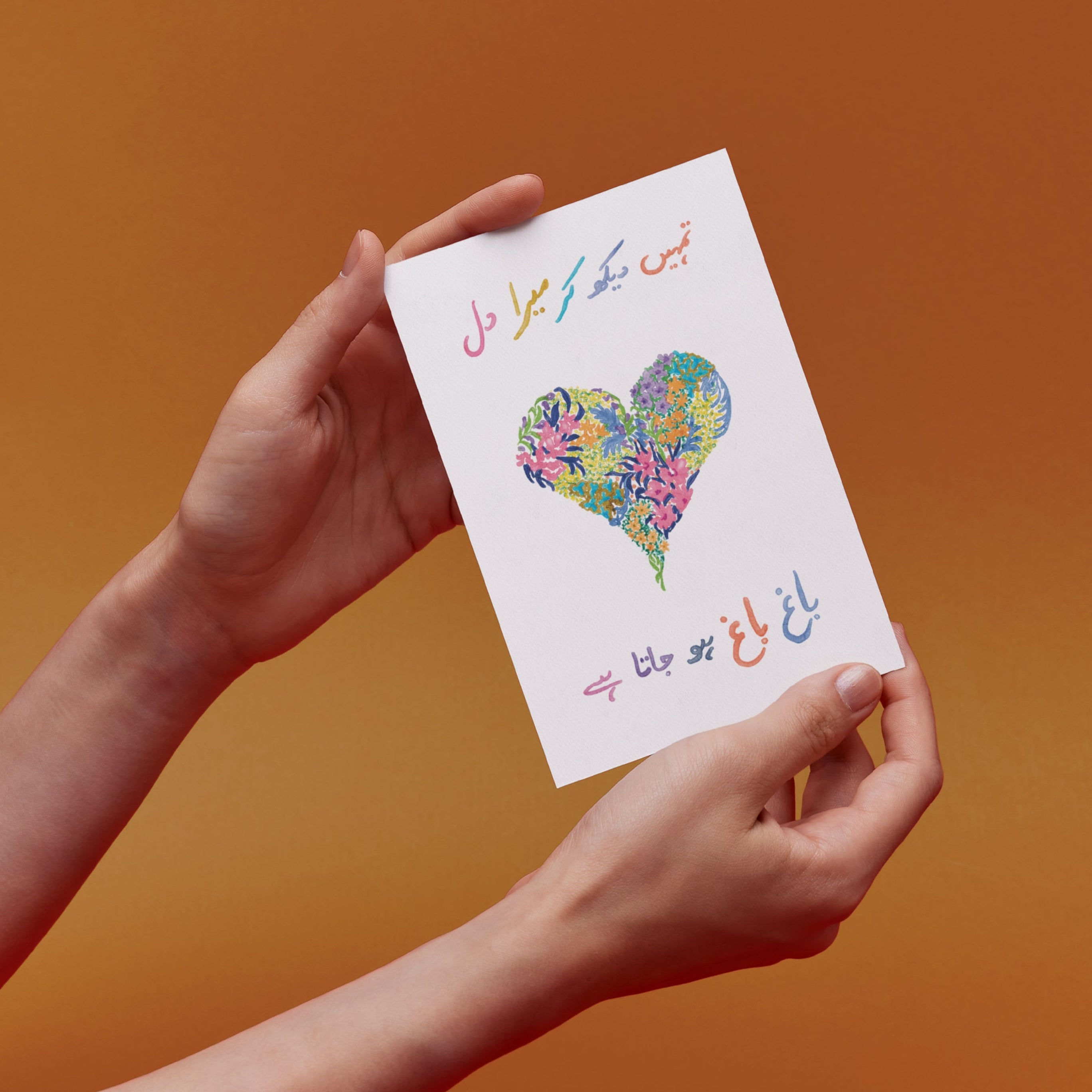 Hands holding a card with an original illustration of a colorful heart design with urdu text translating to "its a joy to see you" 