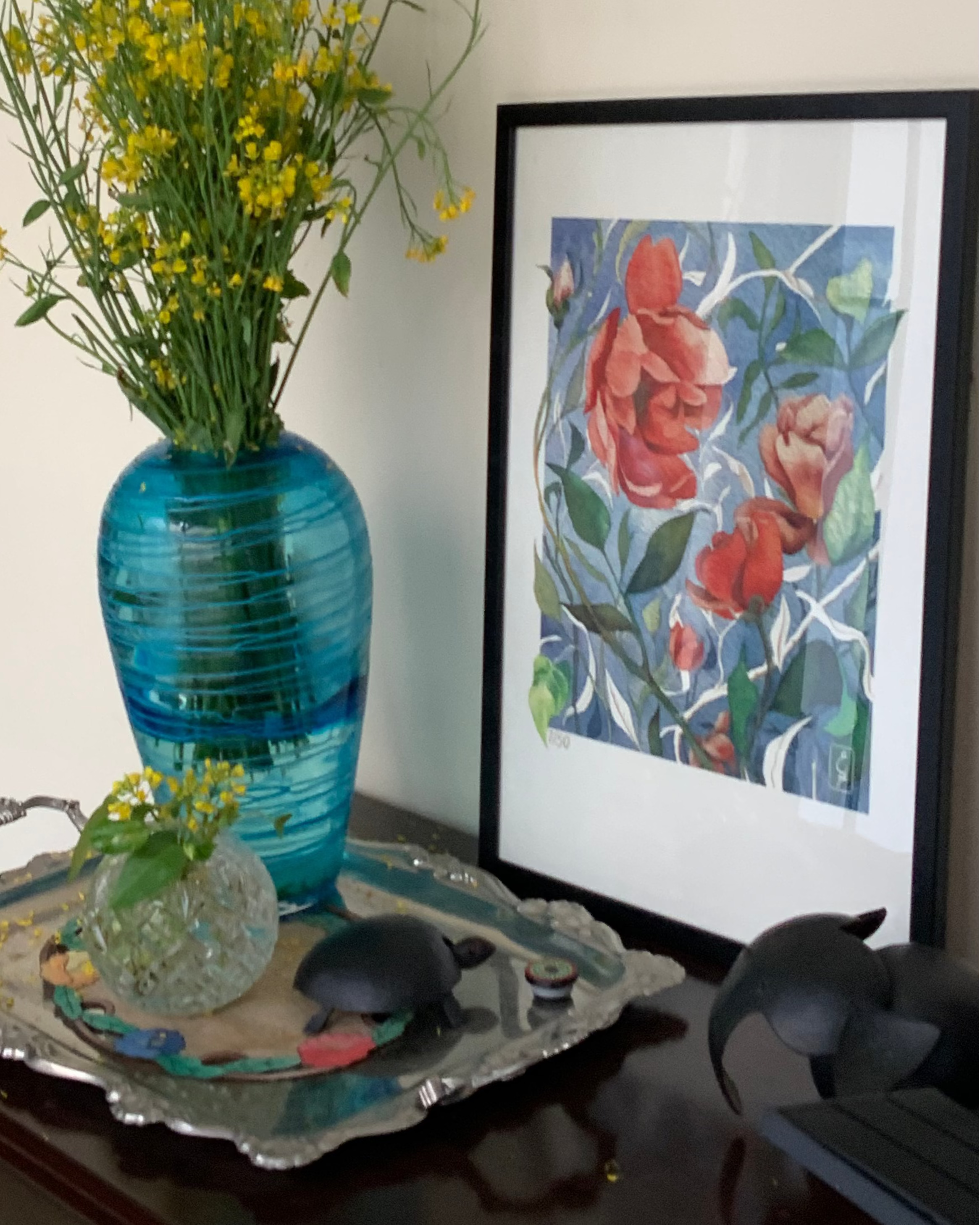 Decorative setting with a blue vase of flowers, silver tray, and floral artwork on a table.