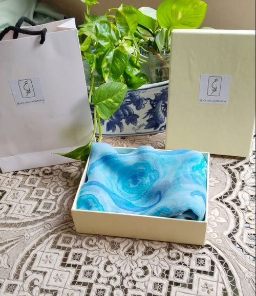 Folded chiffon fabric with hand painted blue swirls, displayed in a box next to Atelier Ambereen packaging 
