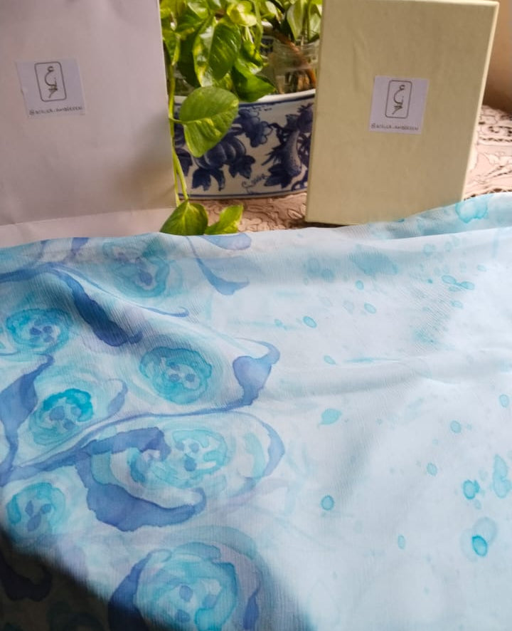 Light blue chiffon fabric draped over a table with visible floral pattern hand painted floral and leaf motifs along the edges. 