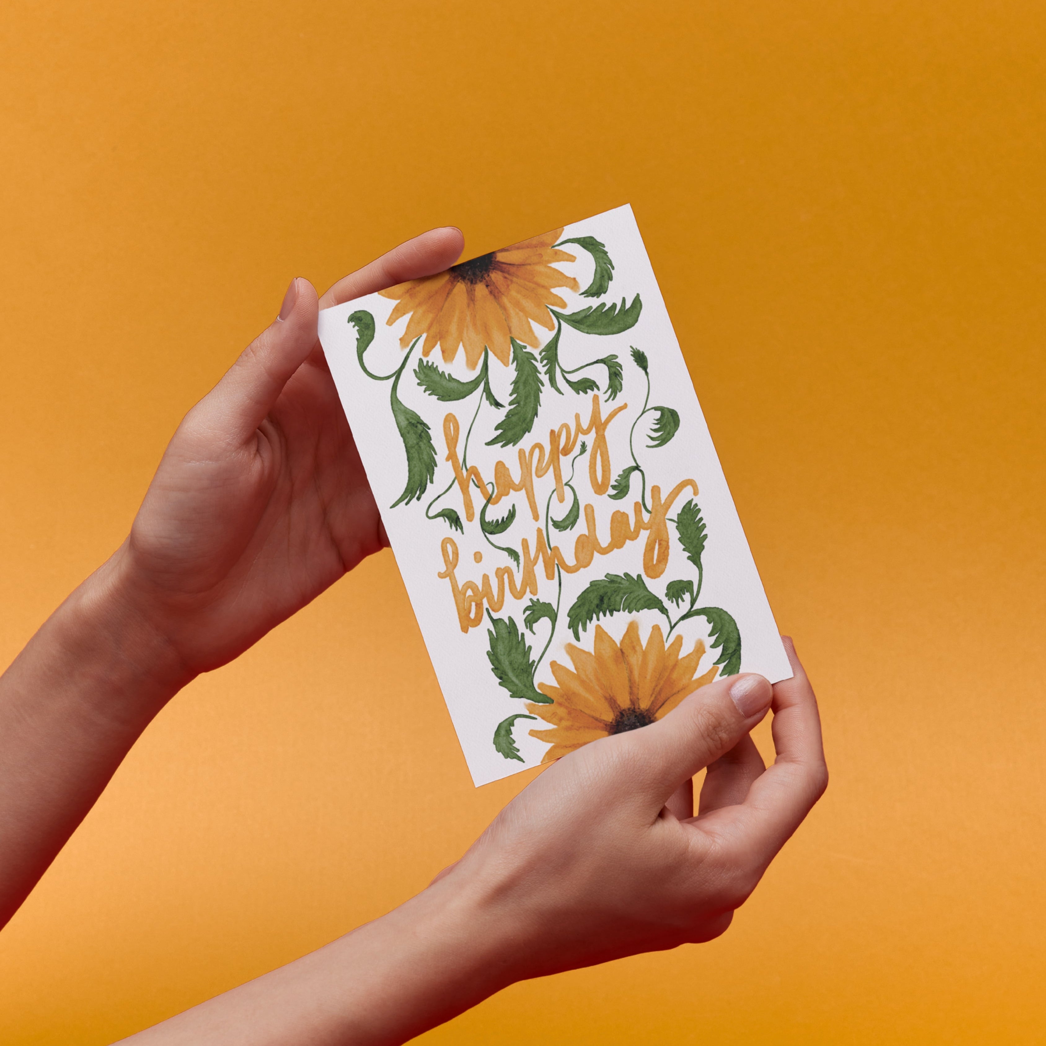 Hand holding a 'Happy Birthday' card with original sunflower design against an orange background