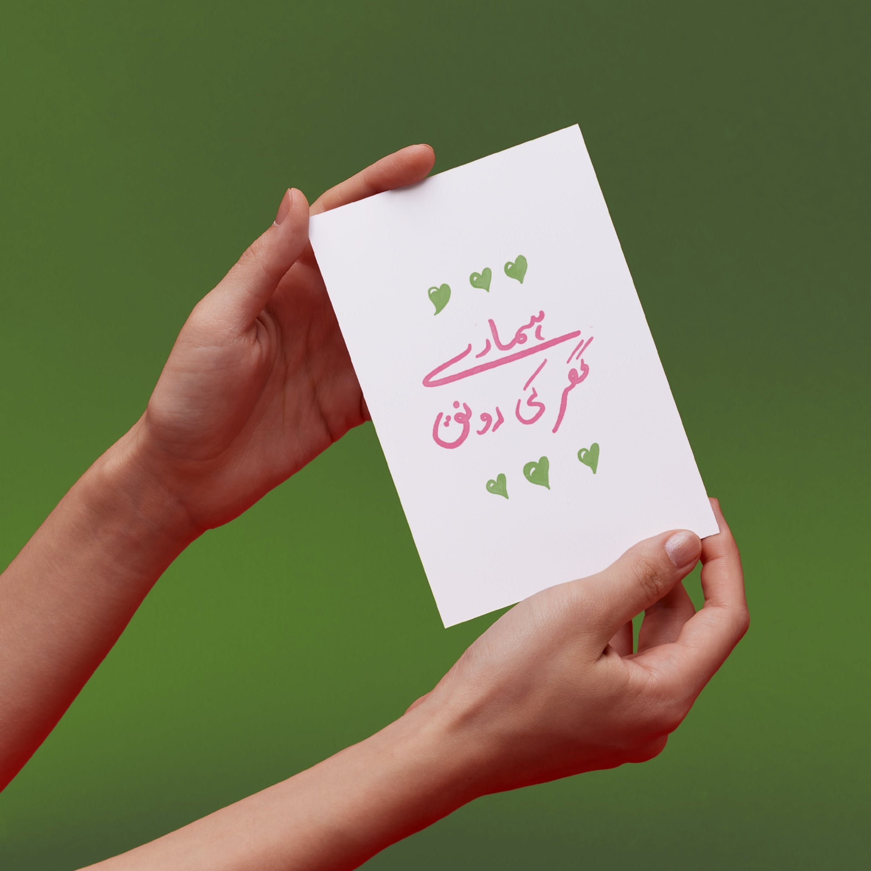 Hands holding up a card with green hearts and pink text in urdu that translates to "The life of our home"