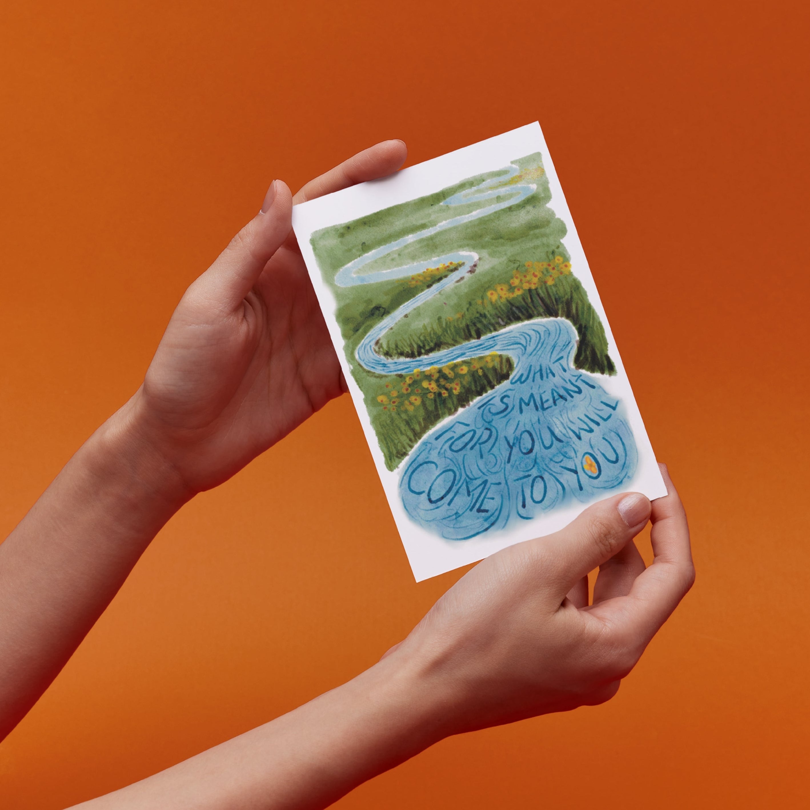 Hands holds a card with a lansdcape illustration of a flower floating in a stream with text saying "What is meant for you will come to you"