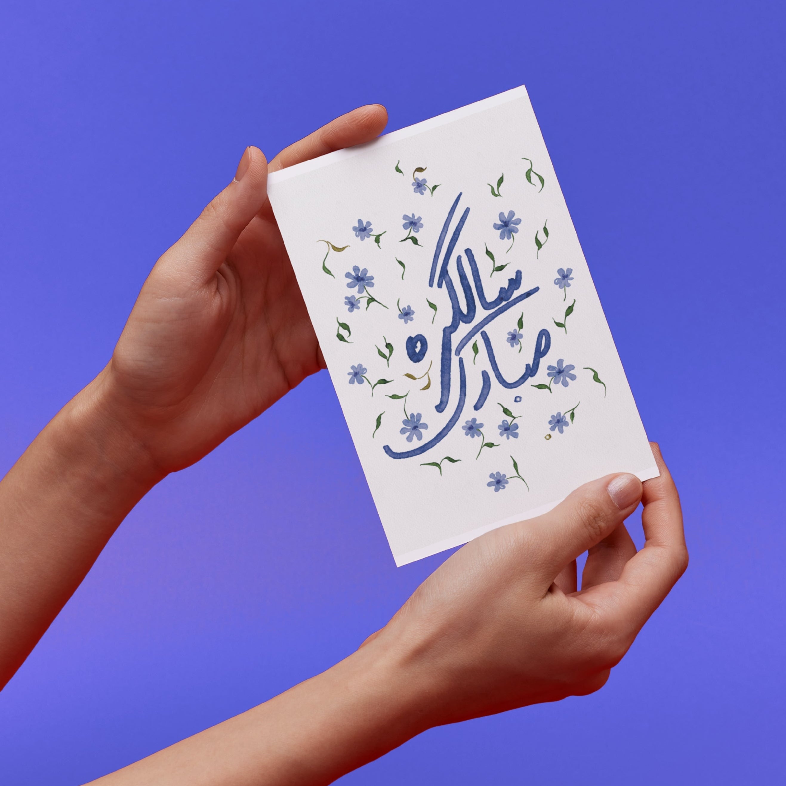 Hands holding a original card design with Happy Birthday in Urdu Text and small floating flowers design against a blue background